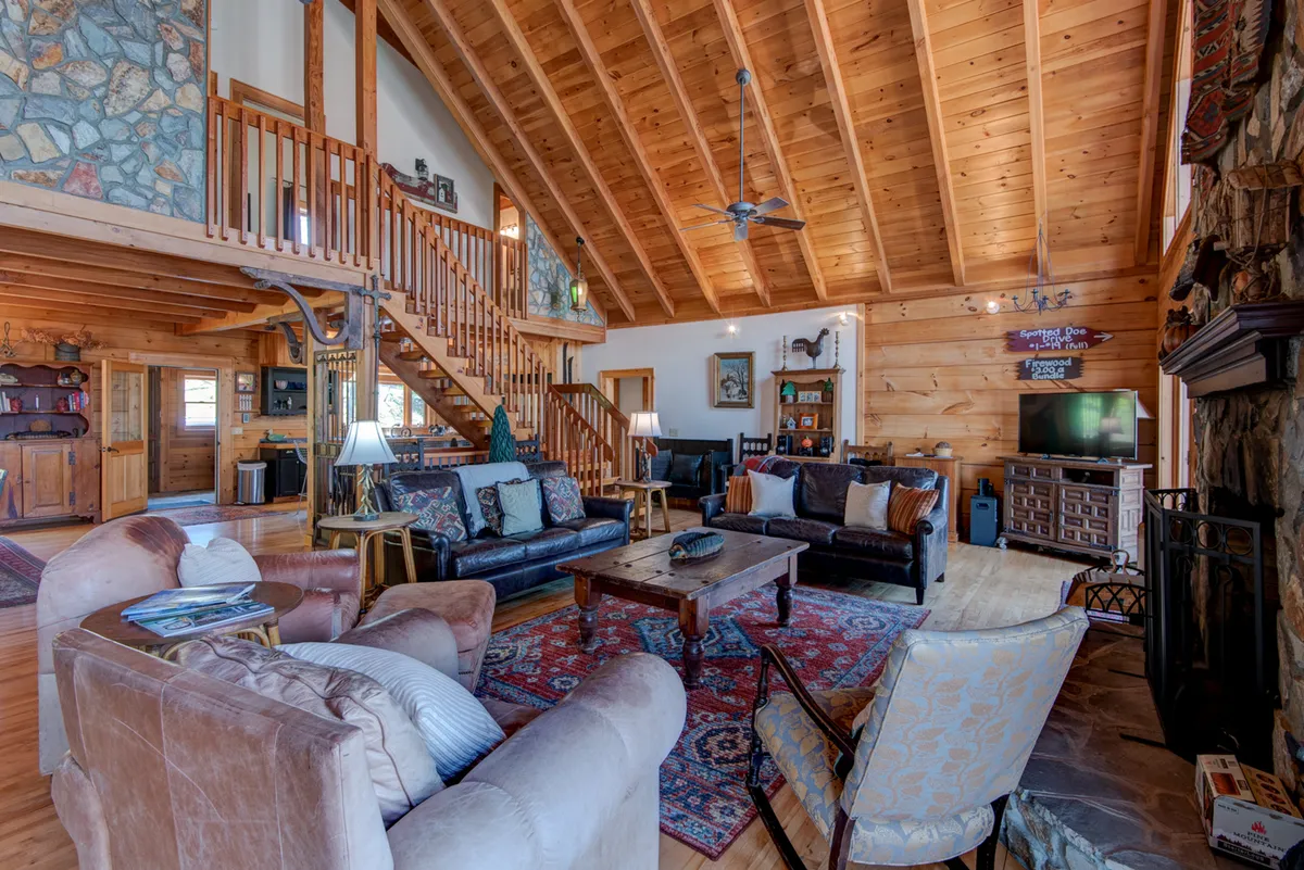 A grand great room with soaring wooden beams, a majestic floor-to-ceiling stone fireplace, and an inviting seating area featuring plush armchairs and a comfortable sofa. Decorative rugs add a cozy touch, and the overall space exudes warmth and togetherness, perfect for family gatherings and shared moments by the fire.