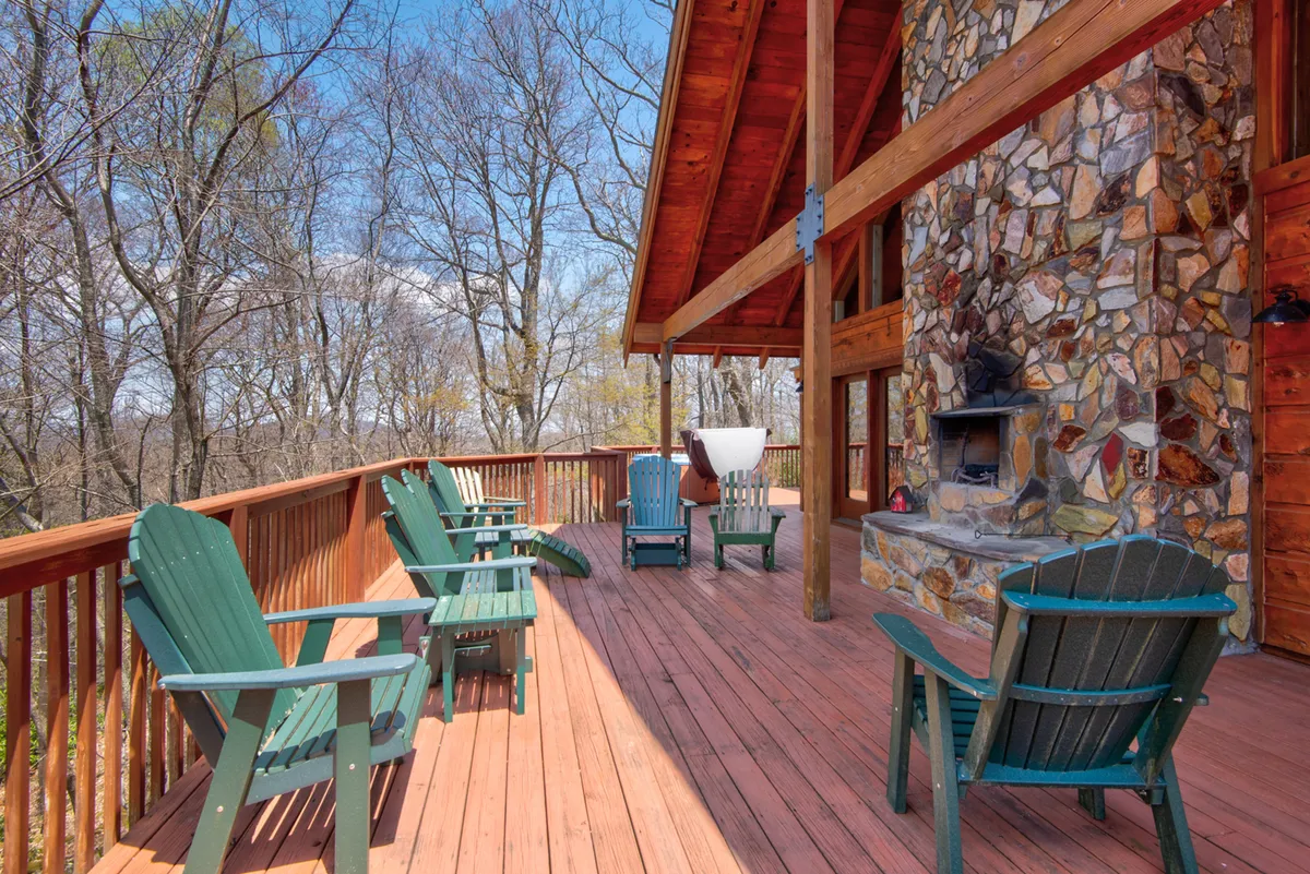A calm, inviting scene of a wooden deck with green chairs arranged near a large stone fireplace. The deck overlooks wooded surroundings, providing a peaceful retreat for gathering around the fire or enjoying the fresh mountain air.