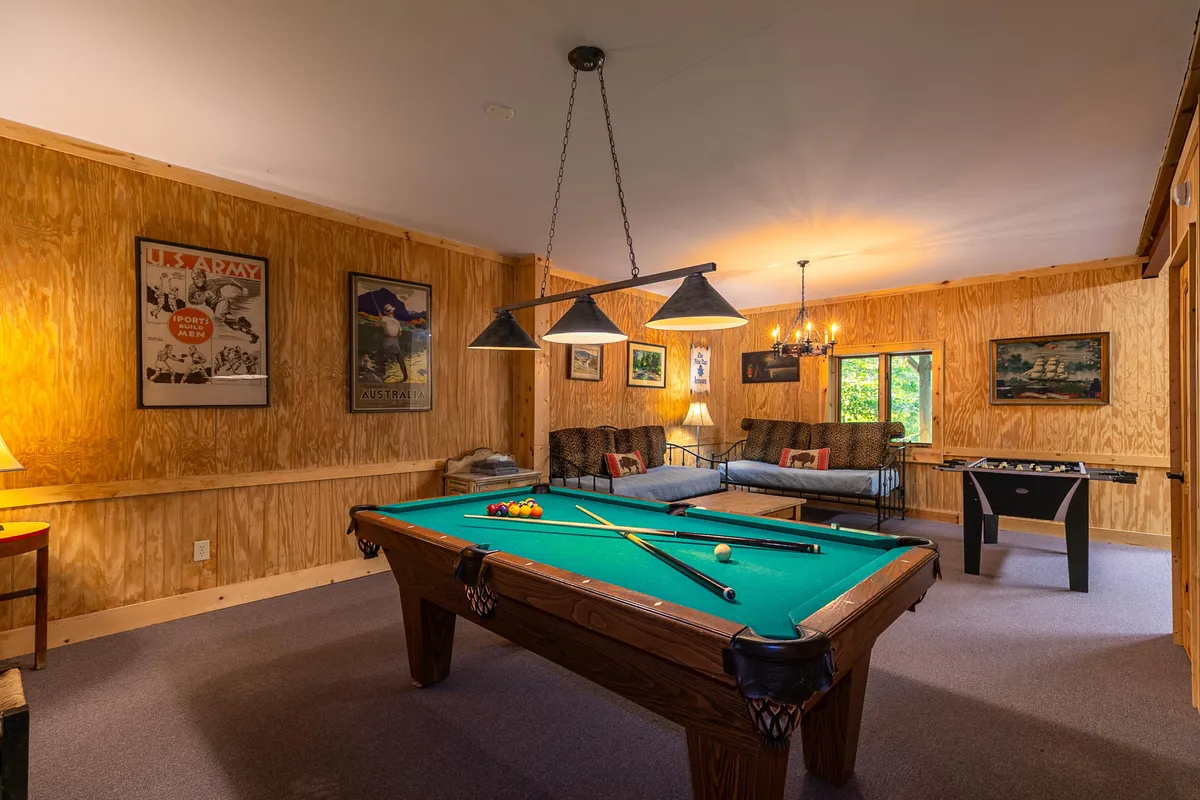 A spacious recreation room with a pool table as the central focal point, surrounded by comfortable sofa seating and striking wall art. Soft lighting enhances the casual, friendly mood, inviting guests to relax, socialize, and enjoy a round of billiards.