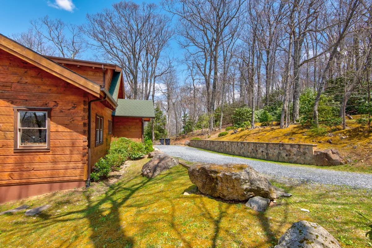The image displays a classic wooden cabin nestled among mature trees, with a gravel pathway curving toward the entrance. Large rocks and dense woods surround the property, suggesting ample space for nature walks, outdoor play, and relaxation. The porch and the wooded acreage reinforce the sense of seclusion and connection to the natural environment.