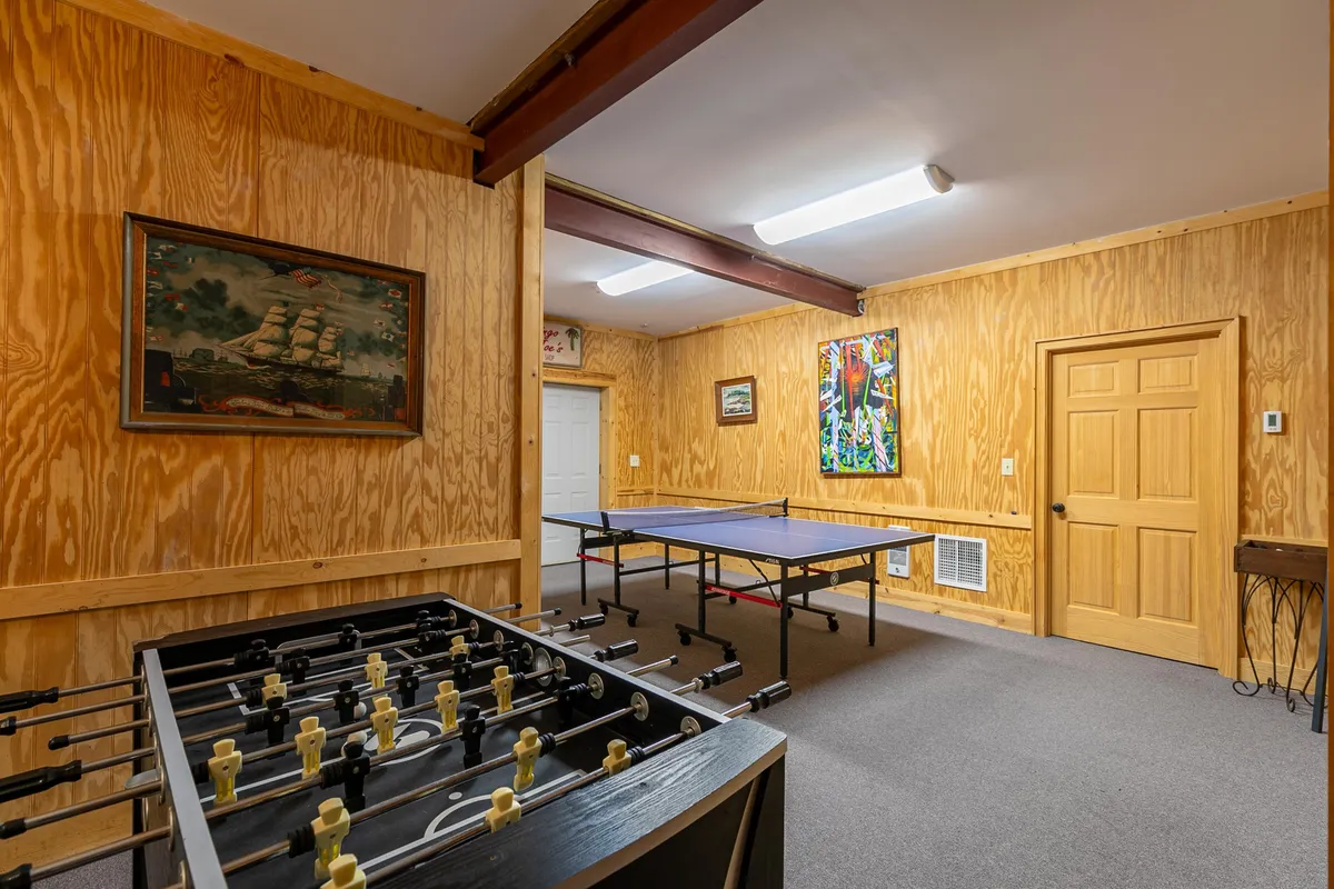 A lively game room scene with a foosball table and a ping pong table set against a backdrop of colorful wall art and a ship painting. The space encourages group play and artistic appreciation, with enough room for multiple activities at once.