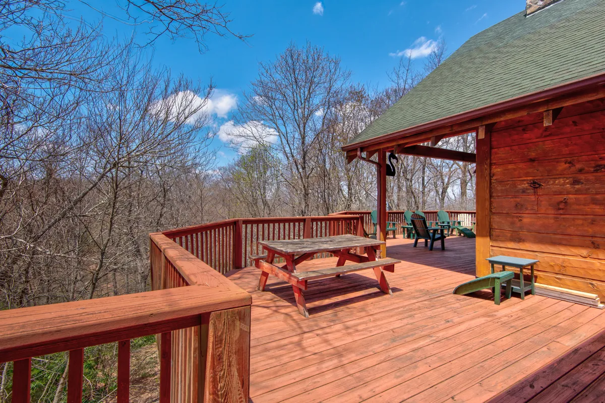 The image features a spacious wooden deck with a picnic table and green chairs, set against a backdrop of trees. The wooden railing and open space invite relaxation, outdoor dining, and enjoying the fresh mountain air. The deck is an ideal spot for lounging with pets or unwinding after a day of exploration.
