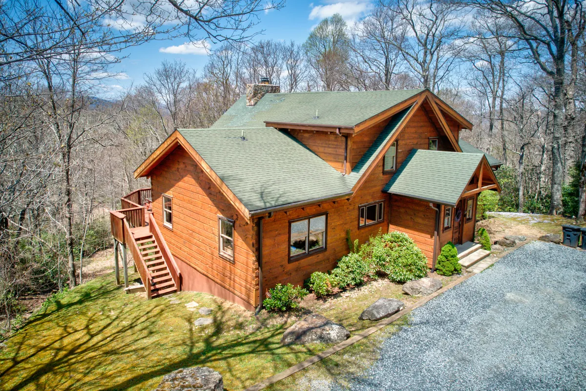 A log cabin structure with a green roof sits amid landscaped gardens and mature trees. The spacious porch invites relaxation, and the surrounding nature hints at plentiful hiking opportunities just steps away. The mood is tranquil and welcoming, ideal for outdoor gatherings.