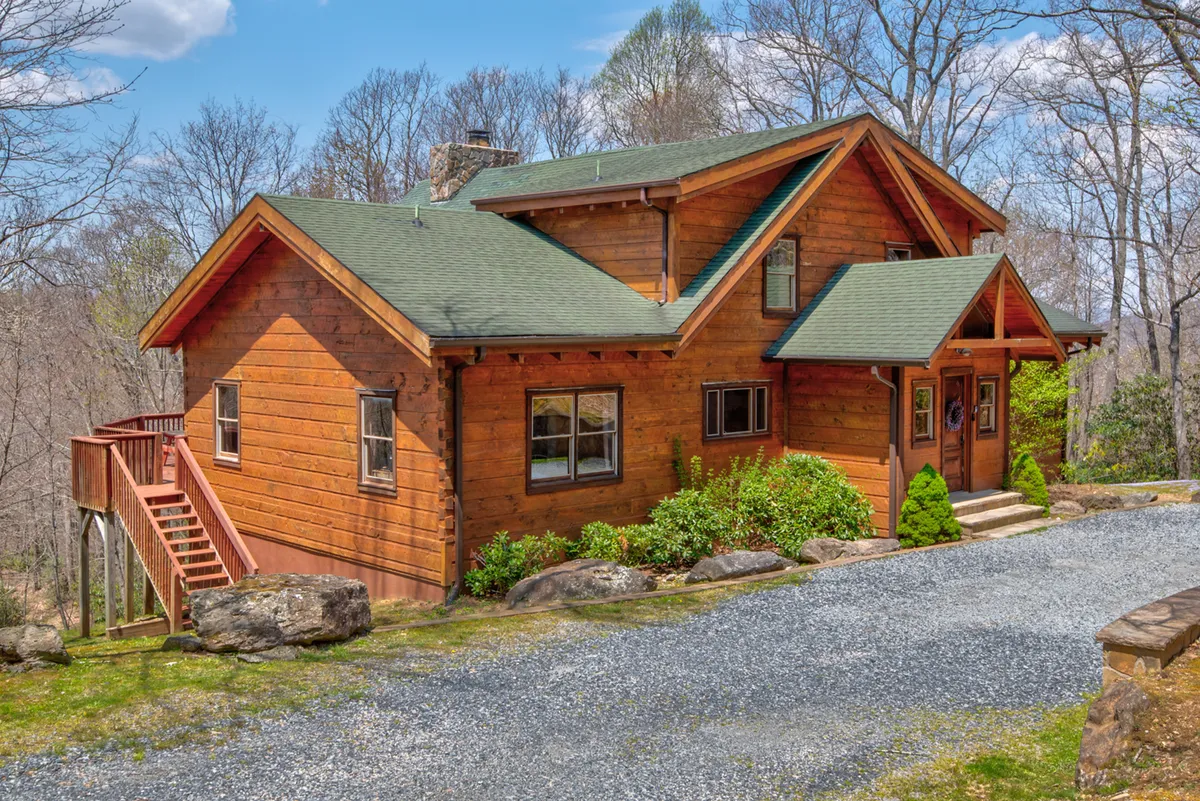 The image showcases the exterior of a spacious wooden cabin with a green roof, nestled in a beautifully landscaped garden. A gravel driveway leads up to the inviting entrance, and the porch offers stunning sunset views. The scene radiates warmth and tranquility, perfect for families or groups seeking a mountain retreat.