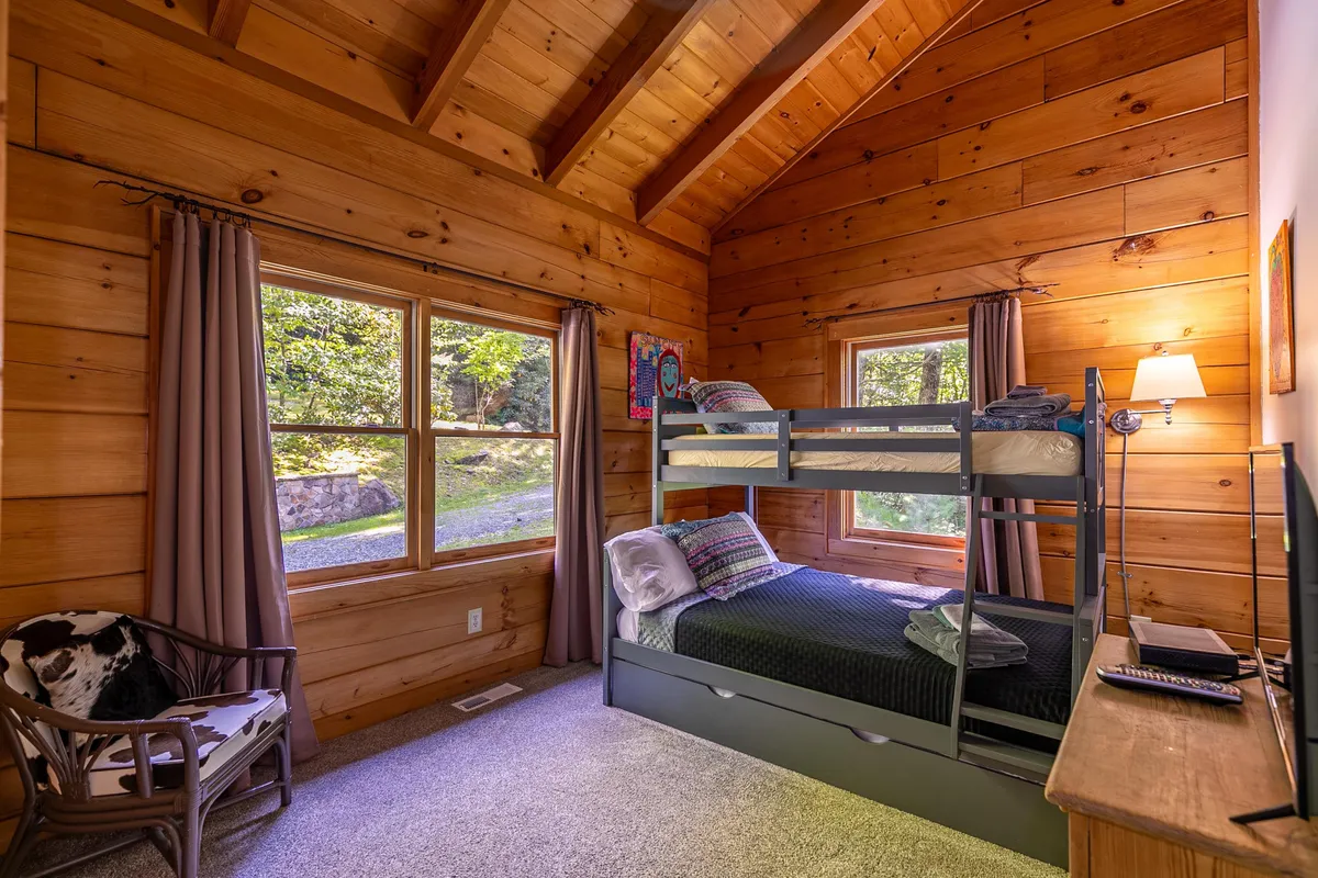 A family-friendly bunk room with a sturdy wooden bunk bed, large windows streaming with sunlight, wooden walls, and an armchair for reading or relaxation. The space is designed for flexibility, accommodating children or extra guests in a cheerful setting.