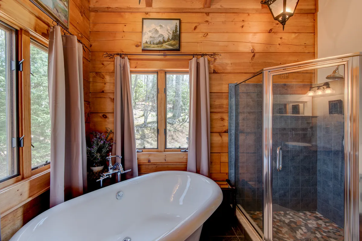 A spa-inspired bathroom featuring a luxurious freestanding bathtub, a glass-enclosed shower, wooden walls, and expansive windows framing views of nature. Decorative artwork and natural light create a serene, rejuvenating environment.