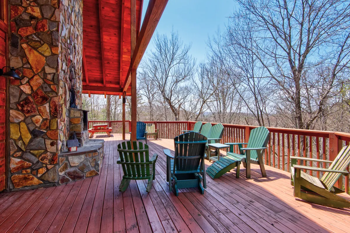 This exterior image showcases a spacious wooden deck with a prominent stone fireplace, surrounded by Adirondack chairs. The backdrop features scenic mountain views, with the warm glow of sunset casting a peaceful ambiance. The space is clearly designed for both relaxation and socializing, with ample room for family gatherings or quiet moments in nature.