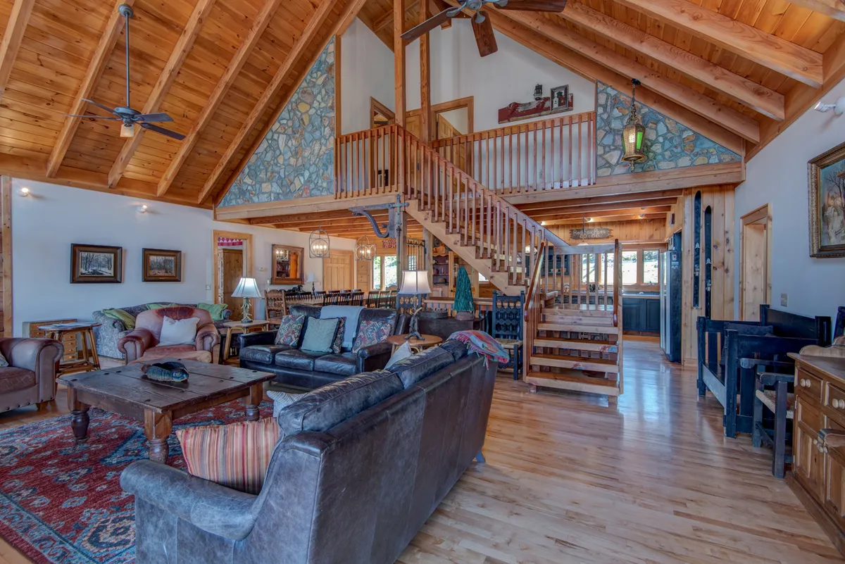 An expansive open-concept living area featuring rustic wooden beams, a dramatic stone fireplace, ample seating with a leather sofa, a nearby staircase leading to the upper floor, and large windows filling the space with natural light. The inviting atmosphere is perfect for socializing, relaxing, and enjoying special moments together.