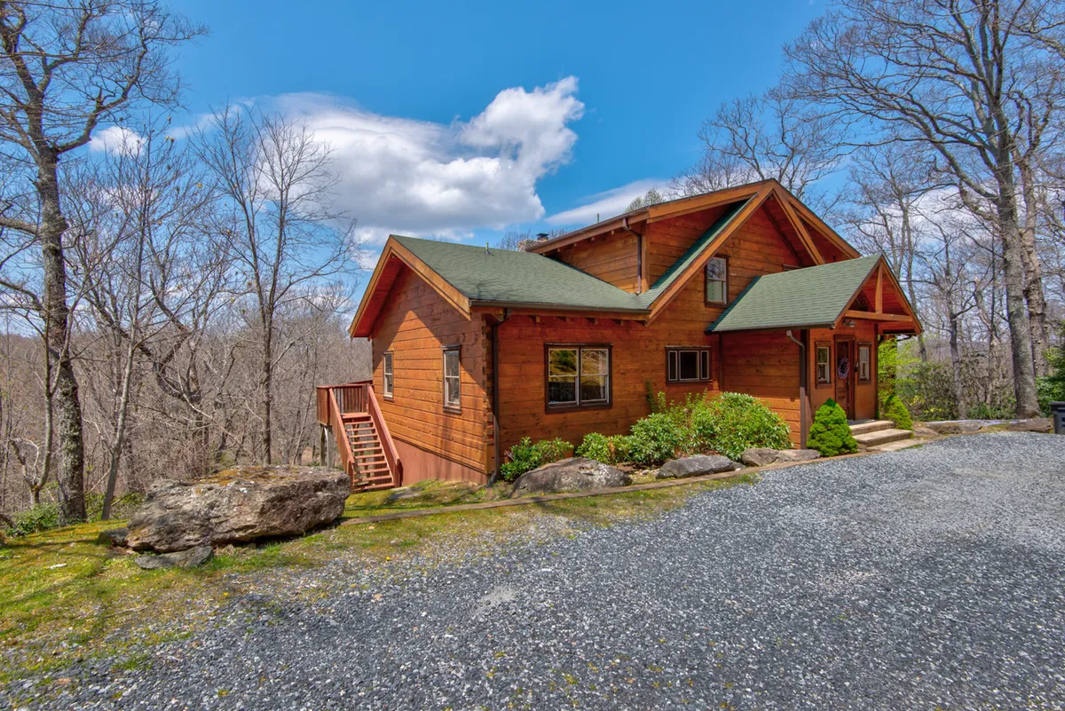 The image features the wooden exterior of Boles Lodge with a green roof, nestled among tall trees. A gravel driveway leads up to the house, framed by natural landscaping. The porch and surrounding forested area suggest a direct connection to the outdoors and easy access to surrounding nature trails.