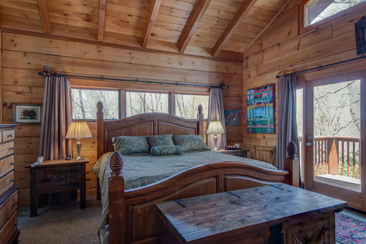 A serene bedroom featuring a wooden bed frame adorned with decorative pillows, a nightstand with a lamp, wall-mounted artwork, and double doors opening out to nature. The space is bathed in soft natural light, creating a tranquil environment ideal for relaxation and privacy.